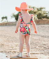 charlarue kids RuffleButts Sunset Garden Ruffle One Piece Swimsuit rear view of ruffles and bow girl on beach in sun hat
